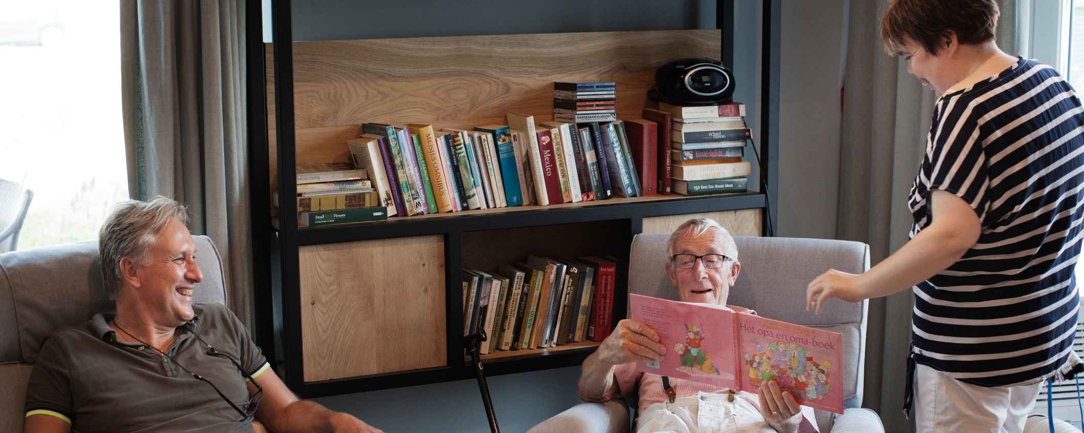 Drie mensen zitten en staan in een woonkamer met stoelen en een boekenkast. Eén persoon bekijkt een boek.