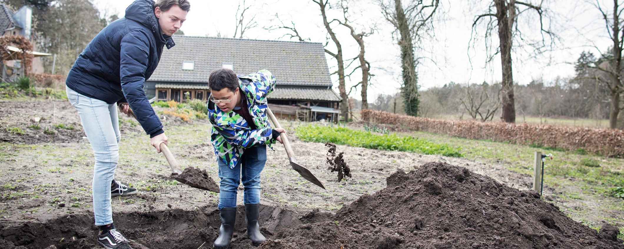 Een volwassene en een kind graven samen in de grond met een schep. Ze werken buiten op een open stuk land bij een gebouw.